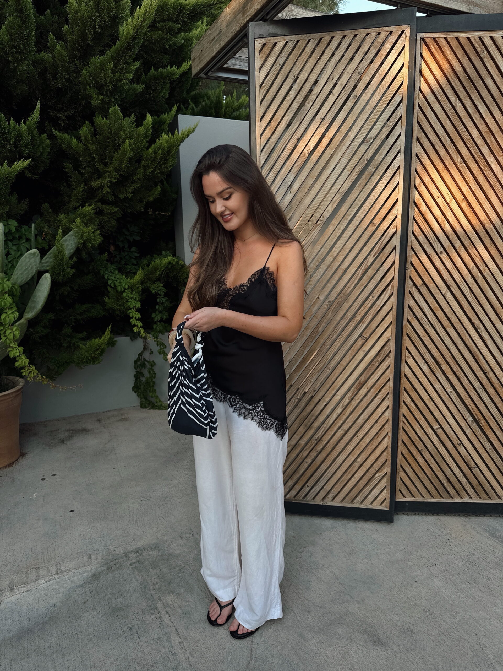 Klasikinė juodai balta dryžuota šilko skarelė – amžinas vasaros akcentas Moteris pozuoja vasaros vakarą, rankoje laikydama klasikinę juodai baltą dryžuotą šilko skarelę – universalų aksesuarą, tinkantį nešioti kaip rankinuką, plaukų aksesuarą ar kaklo puošmeną. Derinys: elegantiškas juodas nėriniuotas satino topas, baltos plačios lininės kelnės, vasariškas stilius, šiuolaikinė miesto mada.